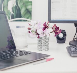 Schreibprozess - Ein Schreibtisch, auf dem zwischen Laptop und Monitor ein kleiner Strauß Blumen in einer Vase steht